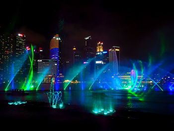 Illuminated modern buildings by river against sky at night