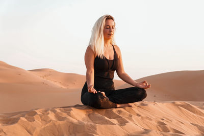 Young woman sitting on sand in desert against sky