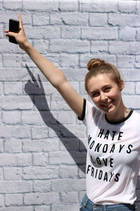 Portrait of smiling teenage girl holding mobile phone while standing against brick wall