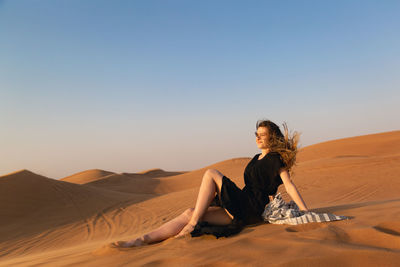 Woman sitting on sand dune in desert against clear sky