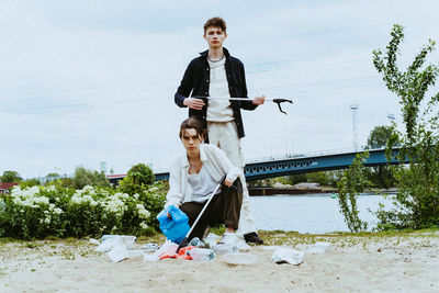 Full length portrait of confident young friends collecting plastic garbage near river against sky