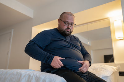 Sad fat man sitting at home bed holds stomach have health problem, insecurities, difficult relations