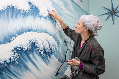 Mature woman artist paints sea waves - mural on a marine theme on the wall