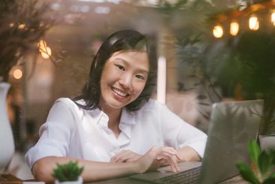 Portrait of happy businesswoman using laptop at table seen through window