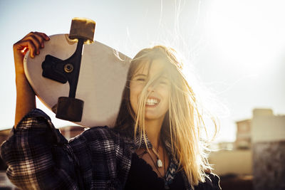 Smiling young woman holding skateboard against sky