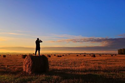 Man standing on hay bale at farm against sky during sunset