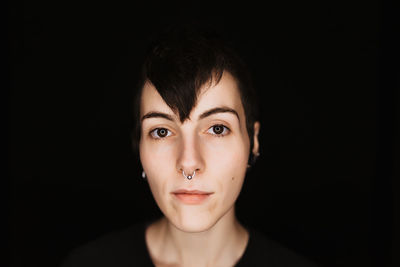 Modern trendy female with short hair and piercing in nose looking at camera against black background