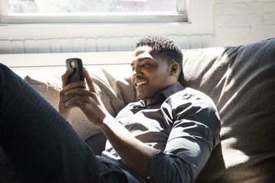 Smiling man using smart phone while lying on sofa at home