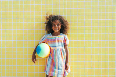 Cute girl with beach ball on sunny day standing in front of wall