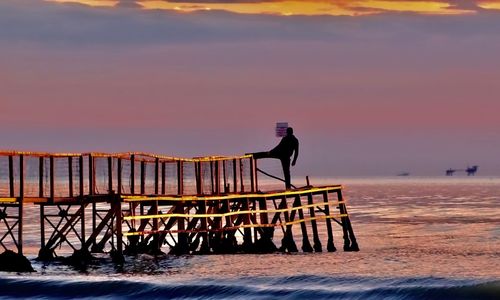 Silhouette man standing on pier at beach against sky during sunset