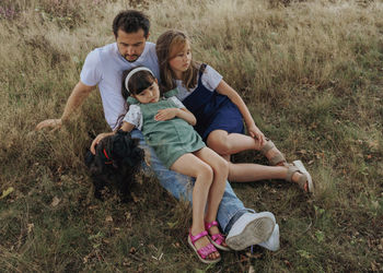 Side view of father with two daughters and little black dog sitting on grassy field