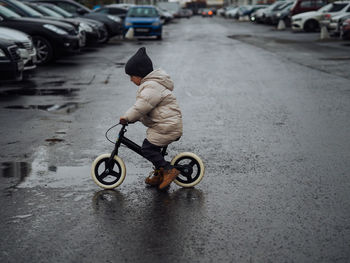 Cute toddler boy in stylish skirt riding on his balance bike crossing a road