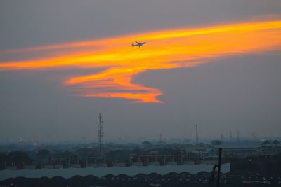 Airplane flying over silhouette cityscape against sky during sunset