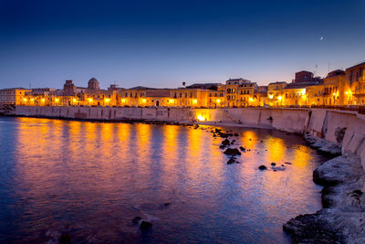 Illuminated buildings by river against sky at night