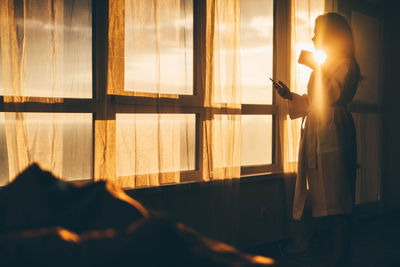 Lady in white bathrobe standing near large window drinking hot coffee in hotel room at sunrise time.