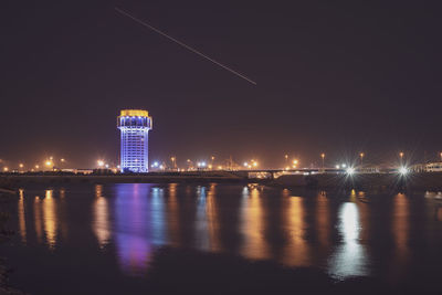 Illuminated buildings by river against sky at night