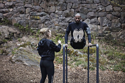 Rear view of female athlete looking at man exercising on parallel bars against stone wall in forest