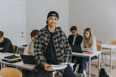 Portrait of smiling young man with book sitting at desk in classroom