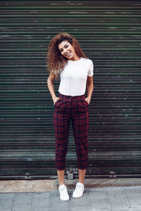 Full length portrait of cheerful woman with hands in pockets standing against shutter