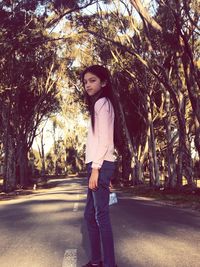 Young woman looking away while standing on road against trees