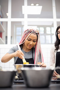 Delighted multiracial women in aprons stirring dish in stainless pot while standing in professional kitchen