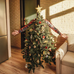 Man with arms outstretched hiding behind christmas tree at home