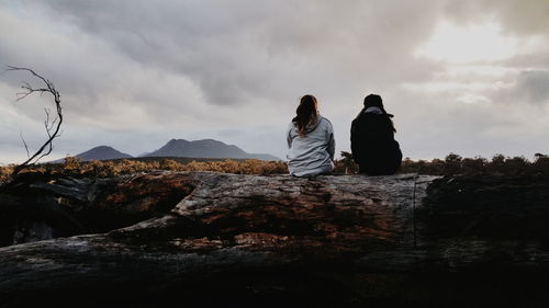 Rear view of friends sitting on rocky beach against cloudy sky