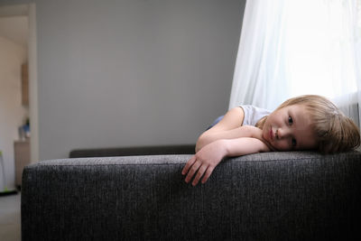 European toddler child sits and gets bored on gray sofa in bright living room, minimalist interior