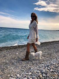 Woman wearing sunglasses with dog walking at beach against sky