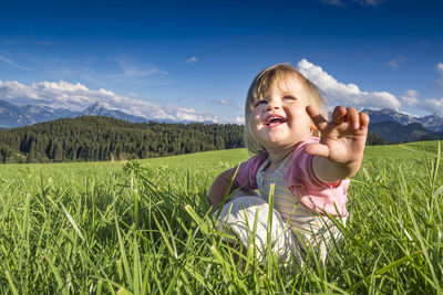 Smiling cute girl gesturing while sitting on grass against sky