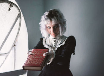 Woman holding book while standing against window at home