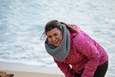 Portrait of smiling young woman wearing warm clothing while bending against sea at beach