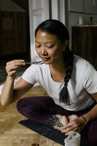 Smiling young woman smelling herbs while sitting on exercise mat at home