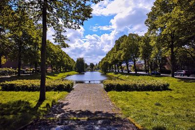 Footpath amidst trees in park with pond against sky