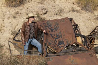 Adult man in cowboy hat with abandoned car in desert, almeria, spain