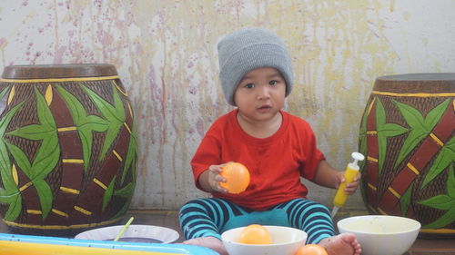 Portrait of cute baby boy holding water bombs while sitting against wall