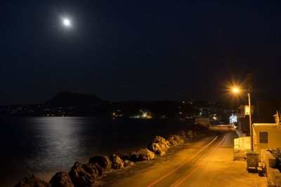 Illuminated street by sea against sky at night