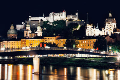 Illuminated bridge over river against buildings at night