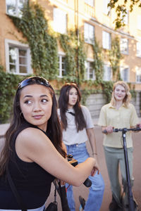 Young female friends spending time together outdoors riding electric scooters