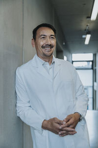 Smiling scientist with hands clasped leaning on wall at clinic corridor