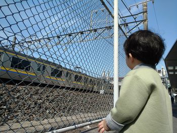 Rear view of boy standing by chainlink fence against clear sky