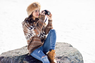 Young woman photographing while sitting on rock during winter