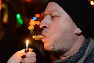 Close-up of man carrying food in mouth while woman hand holding illuminated cigarette lighter at night