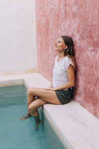 Portrait of young woman sitting leaning against a wall with feet dangling in the pool