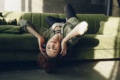 Smiling young woman in headphones lying leaning head down from sofa