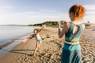Rear view of woman photographing playful daughter standing on shore at beach against sky