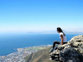 Man sitting on rock by sea against clear blue sky