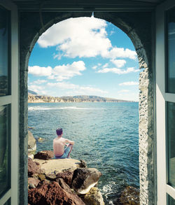 Rear view of shirtless man sitting by sea against sky