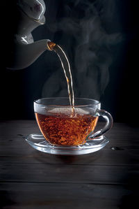 Close-up of teapot pouring drink in cup on table against black background