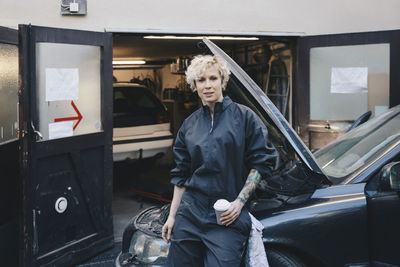 Portrait of confident female mechanic leaning on car outside repair shop